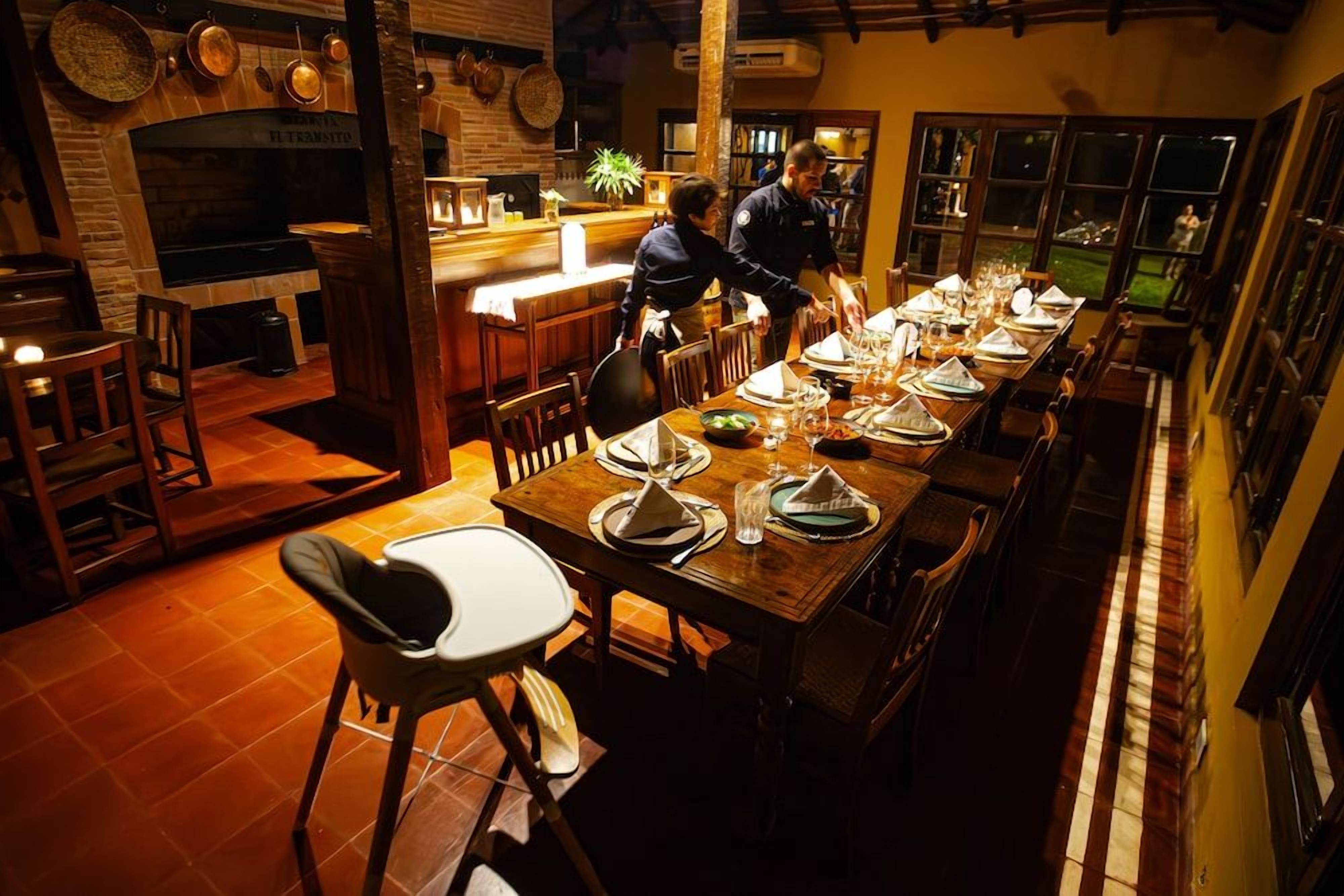 A restaurant with a long dining table set for a meal, a bar area, and a kitchen visible in the background.