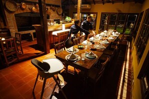 A restaurant with a long dining table set for a meal, a bar area, and a kitchen visible in the background.