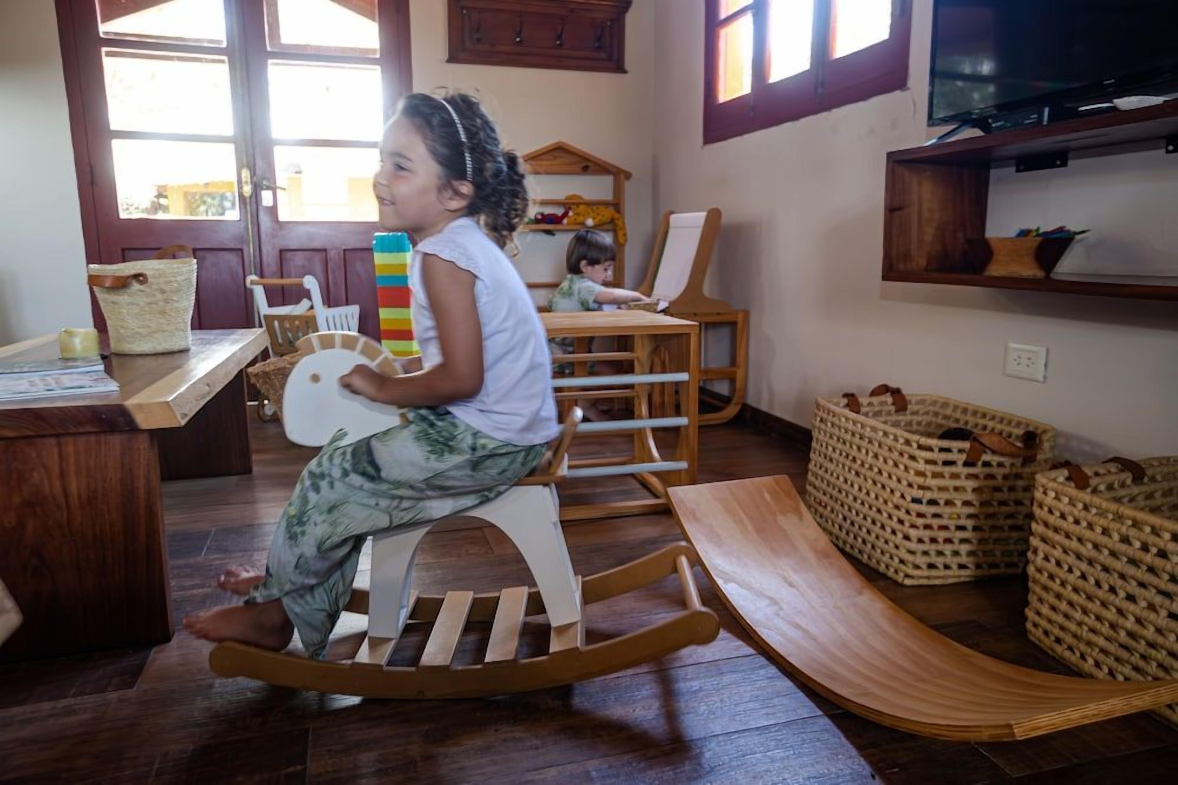 A child on a rocking chair in a room with a wooden floor, a desk with a basket, and a television on the wall.