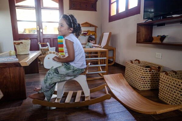 A child on a rocking chair in a room with a wooden floor, a desk with a basket, and a television on the wall.