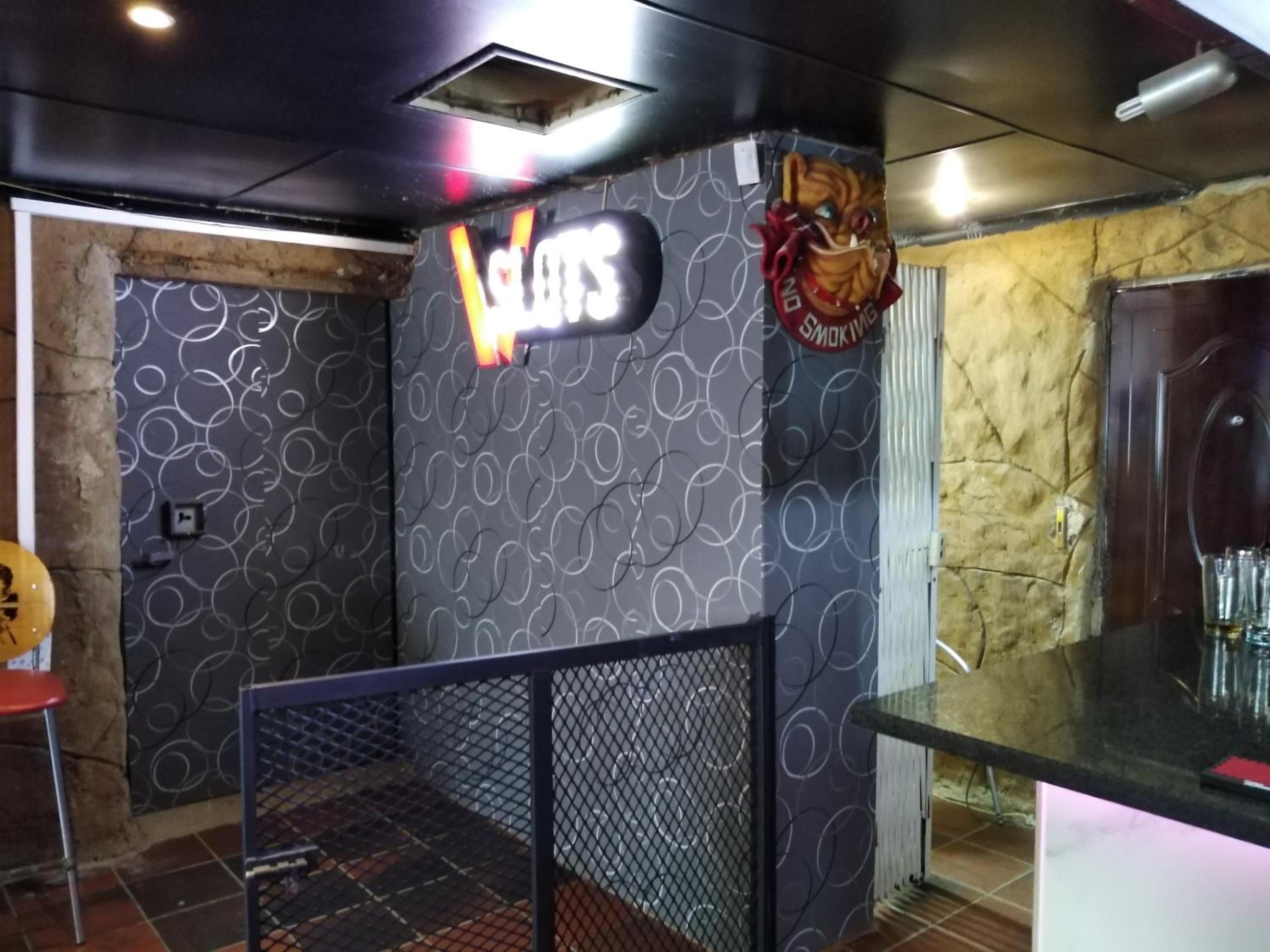 A room with a patterned wall, a sign reading 'Slots', a decorative mask, and a bar area with glasses.