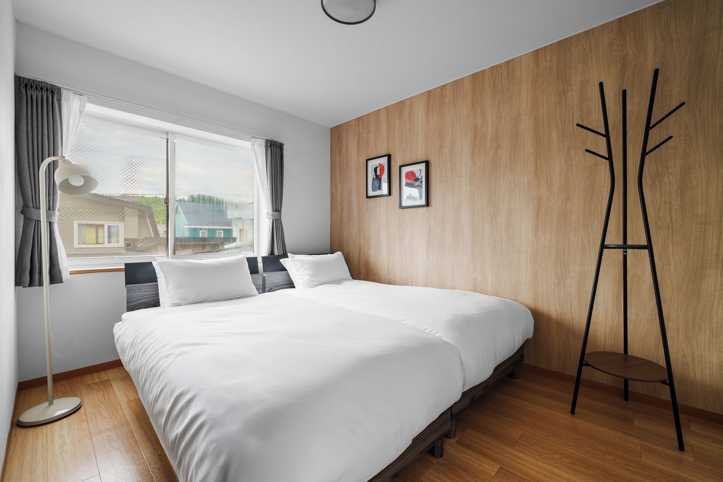 A bedroom with a large bed, a standing lamp, a window with curtains, and two framed pictures on the wall.