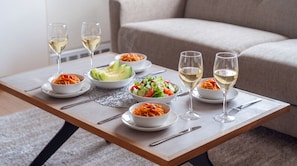 Uma mesa de jantar com diversos pratos e copos de vinho branco, sobre um tapete, com um sofá ao fundo.