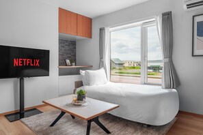Quarto de hotel moderno com cama, televisão fixada na parede, uma pequena mesa com uma planta e janela com cortinas.