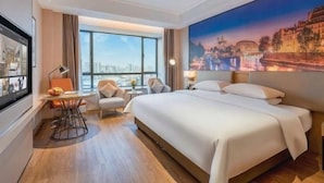 Hotellihuone, jossa on suuri sänky, työpöytä tuolilla, ikkuna, josta avautuu näkymä kaupunkiin, ja seinällä oleva taulu.