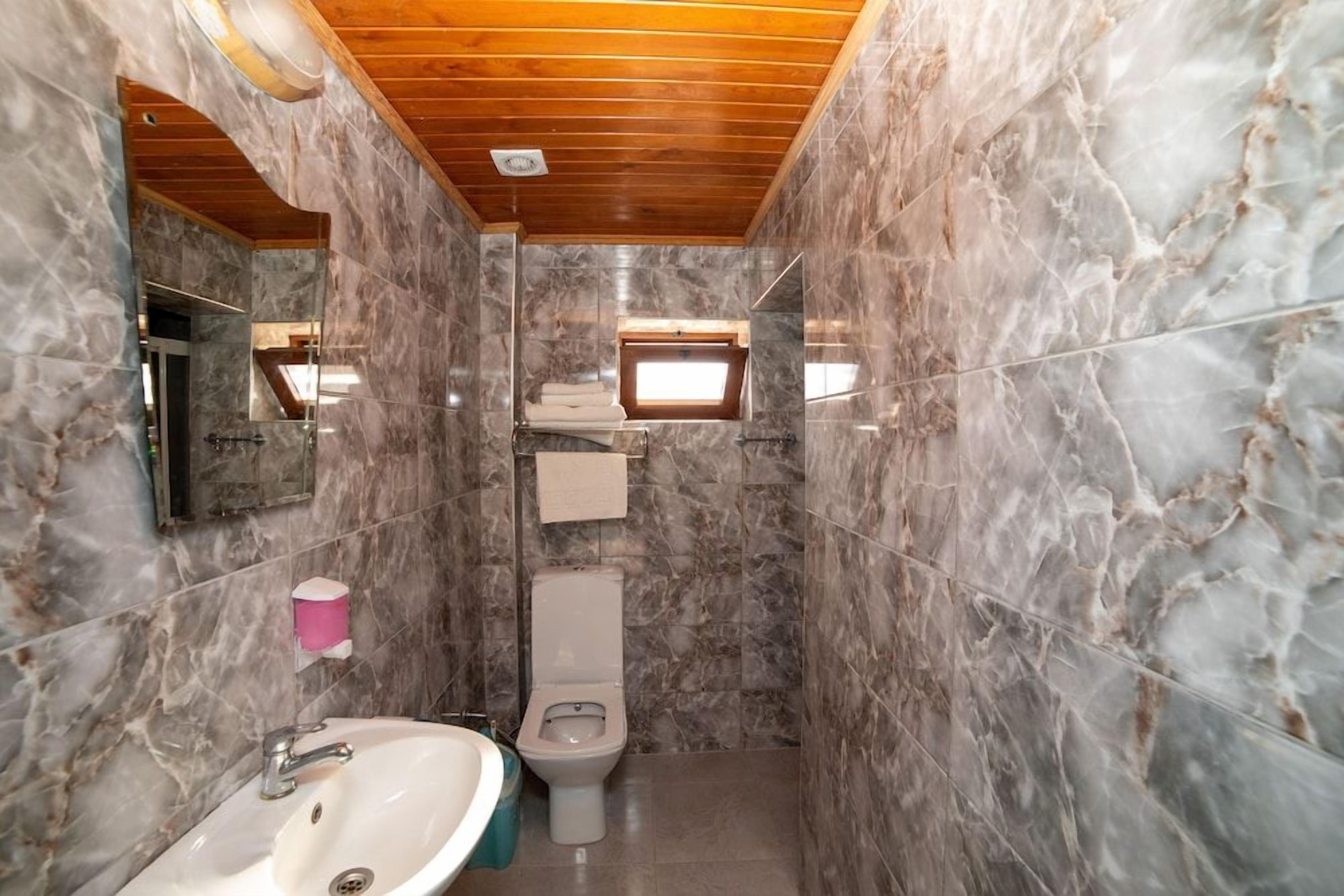 A bathroom with marble walls, a wooden ceiling, a sink, a toilet, and a window.