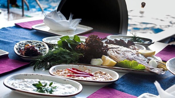 Mesa de jantar com diversos pratos, piscina ao fundo e um guarda-sol preto.