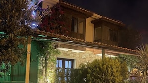 Una casa con fachada de piedra, un porche cubierto y un jardín bien iluminado durante la noche.
