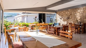 Un área de comedor al aire libre con varias mesas y sillas de madera, una pared de piedra y vistas a edificios al fondo.