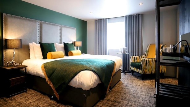 A hotel room with a large bed, two bedside tables with lamps, a chair, a desk with a chair, and a view of the outdoors. - Deira Hotel & Apartments (Prestwick)