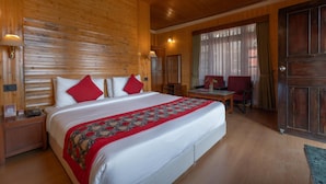 Quarto de hotel com uma cama grande, uma escrivaninha e uma cadeira. É visível uma parede de madeira e uma porta.