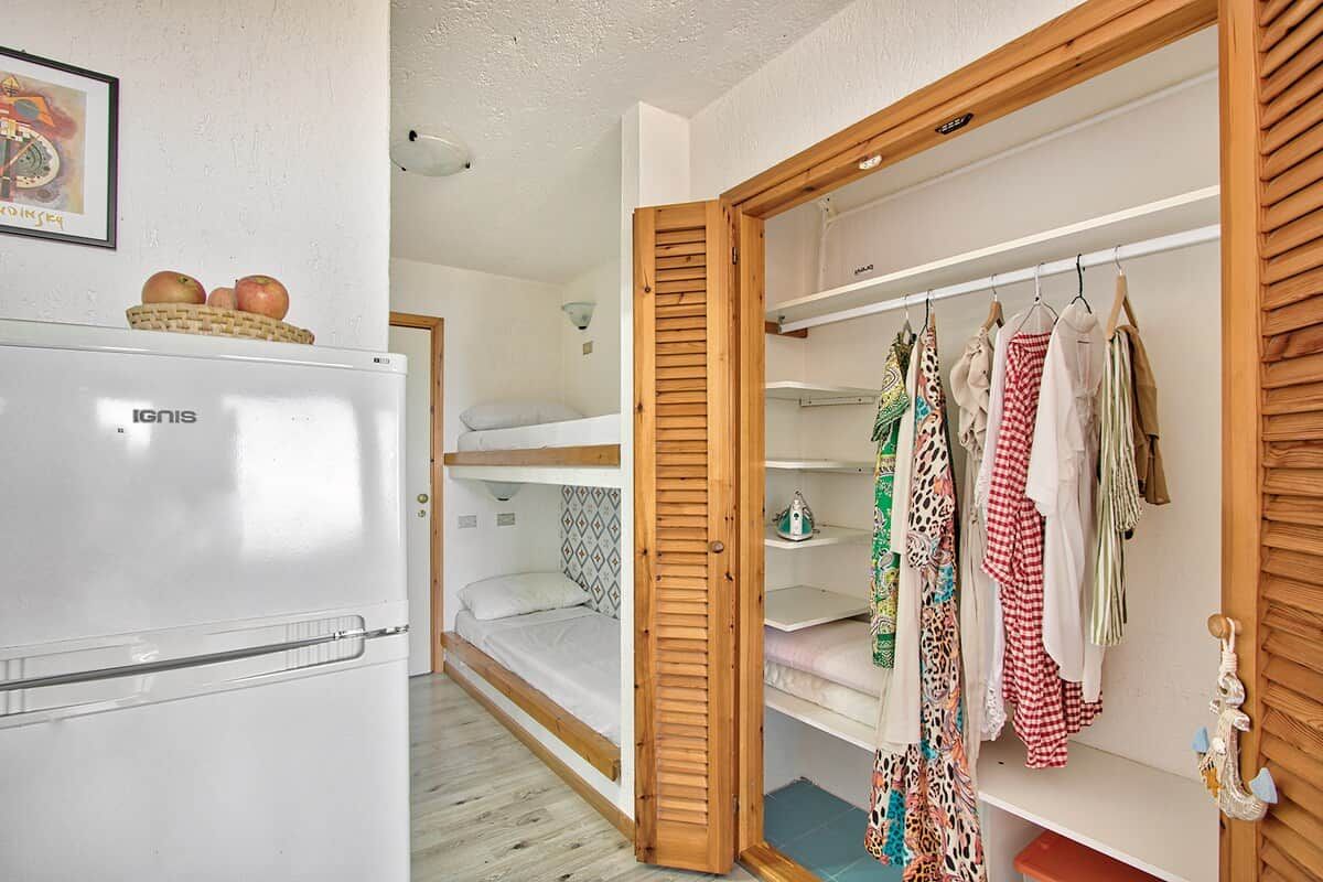 Quarto compacto com beliches, guarda-roupa com roupas penduradas e um frigorífico.