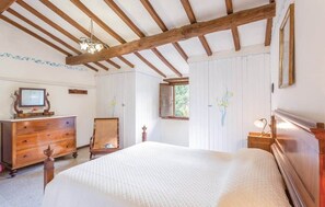 Quarto com teto de madeira, cama com colcha branca, cômoda, cadeira, televisão e janela com vista para vegetação.