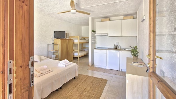 Een kleine slaapkamer met een bed, nachtkastje en plafondventilator. Grenzend daar aan bevindt zich een kitchenette met een spoelbak, fornuis en kastjes.