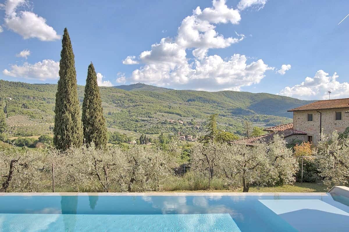 A pool with a view of a mountainous landscape, two tall cypress trees, and a building with a tiled roof.