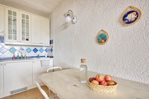 A kitchen with white cabinets, a tiled backsplash, a table with a basket of fruit, and wall-mounted decorative items. - Comfort and relaxation in the heart of the Costa Smeralda (Cugnana Verde)