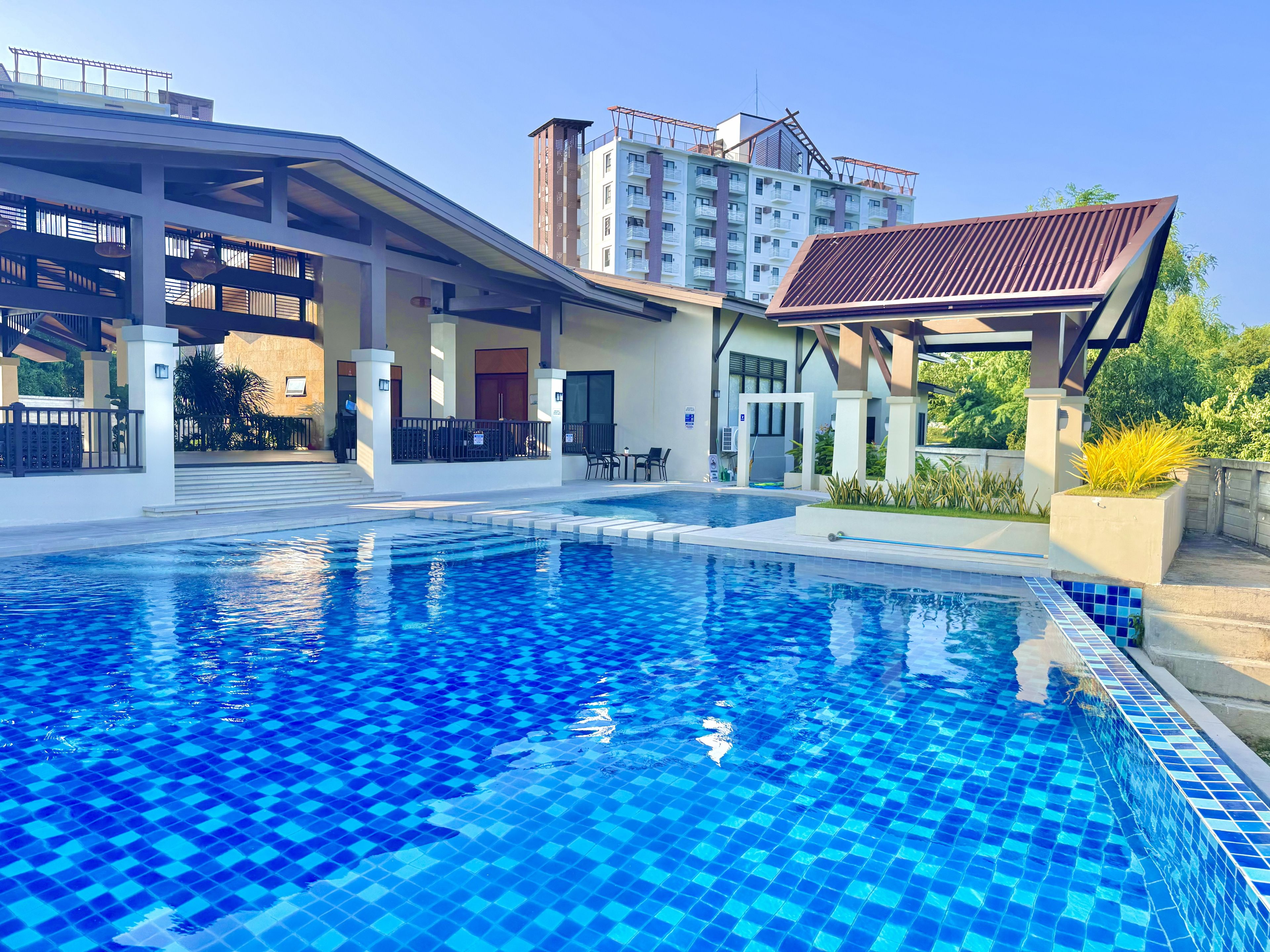 A swimming pool with a tiled deck, a covered seating area, and a building with multiple floors in the background.