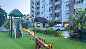 A playground with seesaw and climbing equipment, surrounded by a well-maintained lawn and a residential building with balconies. - Royal Oceancrest Panglao 1 (Dauis)