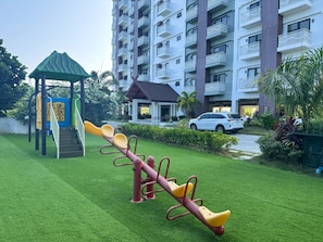 A playground with seesaw and climbing equipment, surrounded by a well-maintained lawn and a residential building with balconies. - Royal Oceancrest Panglao 1 (Dauis)