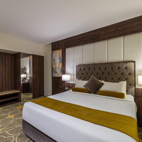 A hotel room with a large bed, a bedside table, a lamp, a mirror, and a wooden wardrobe. - Gardn Millennium Hotel Abha (Abha)