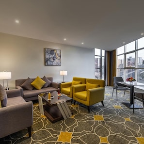 Moderni hotellihuone, jossa on sohva, nojatuoleja, sohvapöytä, ruokailutila ja suuri ikkuna, josta avautuu näkymä kaupunkiin.