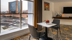 A hotel room with a large window, a flat-screen TV, a dining table with chairs, and a fruit bowl. - Gardn Millennium Hotel Abha (Abha)