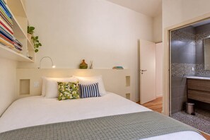 A bedroom with a bed, pillows, a bedside table, a wall-mounted shelf with books, and a door leading to a bathroom. - Villa Surf House Plage de l'Horizon et commerces a pied (Lège-Cap-Ferret)