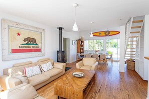 A spacious living area with a sofa, coffee table, and a dining table set for four. A large framed picture of a bear is on the wall, and a surfboard hangs from the ceiling. - Villa Surf House Plage de l'Horizon et commerces a pied (Lège-Cap-Ferret)