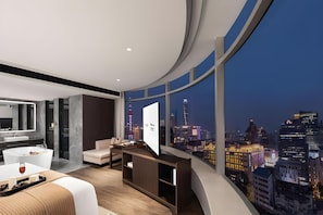 Een moderne hotelkamer met een gebogen balkon, uitzicht over de stad, een flatscreen televisie, een bank, een eettafel en een grote spiegel.
