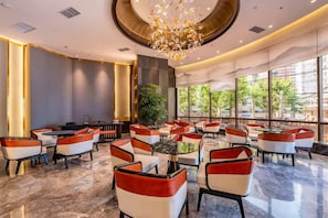 Een hotellobby met een modern design, met een centraal zitgedeelte met ronde tafels en stoelen, een grote kroonluchter en grote ramen met uitzicht op het exterieur.
