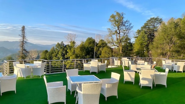 An outdoor seating area with white wicker furniture and a green lawn, overlooking a mountainous landscape. - Pinemont Hotel Murree (Murree)
