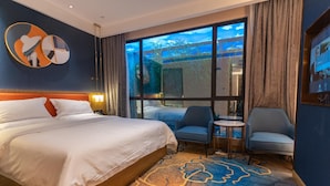 Habitación de hotel con una cama grande, dos sillones, una mesita y vistas a un paisaje tropical a través de las ventanas.