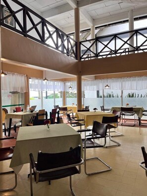 Un vaste espace intérieur aménagé en salle à manger, avec de nombreuses tables et chaises, de grandes fenêtres et un plafond haut avec des poutres apparentes.