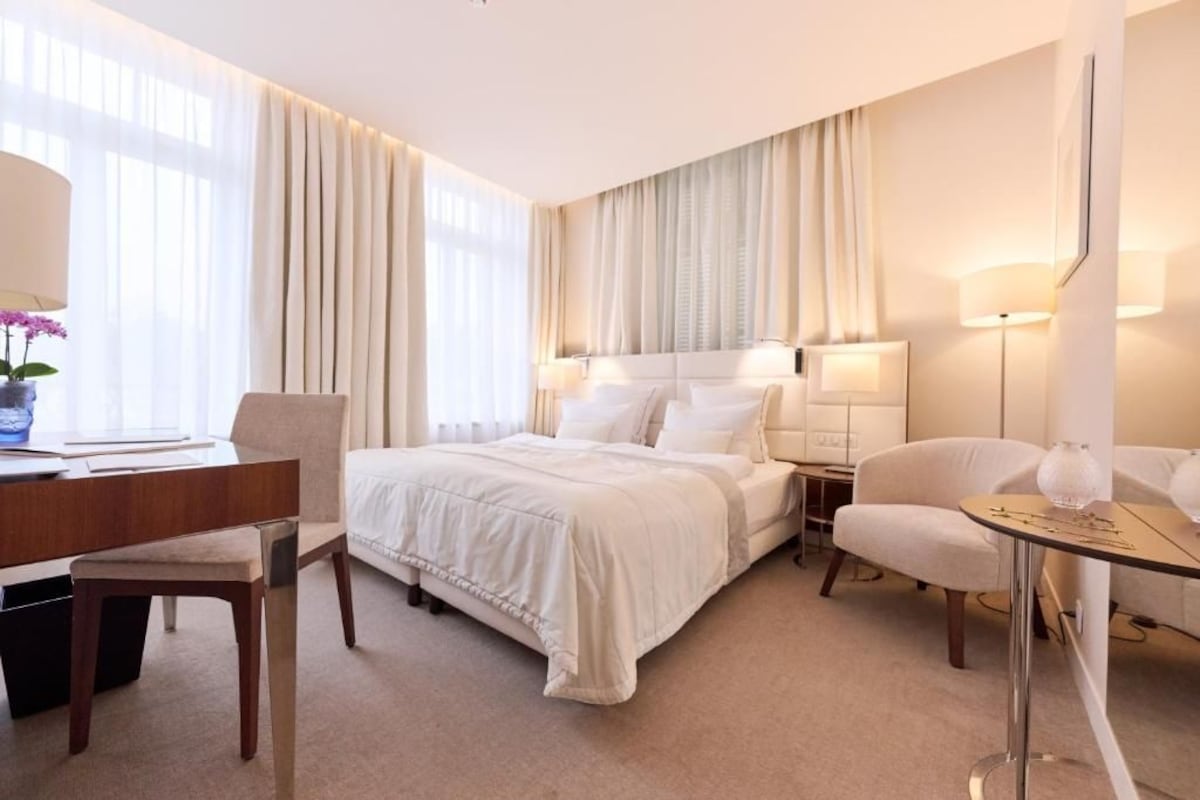 A hotel room with a bed, bedside tables, a desk, a chair, and a small table with a lamp.