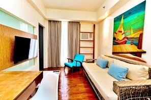 Una camera d'albergo con un divano, una poltrona, una televisione, un quadro e una finestra con tende.