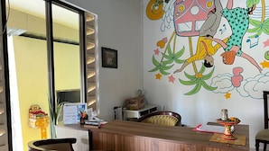 A reception desk with a nameplate, a window with blinds, and a colorful mural on the wall. - Collection O Near Monkey Forest Formerly Hepiyuk Guest House (Ubud)