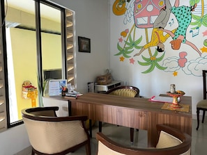 A reception desk with a nameplate, a window with blinds, and a colorful mural on the wall. - Collection O Near Monkey Forest Formerly Hepiyuk Guest House (Ubud)