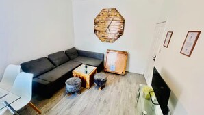 A modern living room with a grey sectional sofa, a wooden octagonal clock, a small wooden cabinet, a glass coffee table, and a TV stand. - 4-bed House, Sleeps 9 w Garden & On-street Parking (Liverpool)