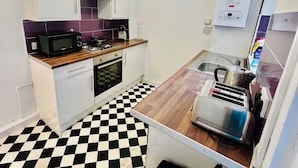 A compact kitchen with white cabinets, a microwave, a gas stove, a toaster, and a checkered floor. - 4-bed House, Sleeps 9 w Garden & On-street Parking (Liverpool)