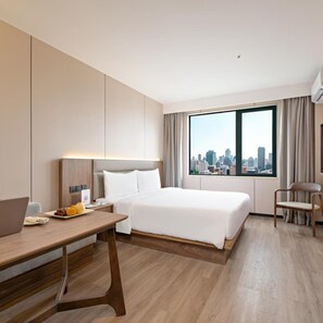 Quarto de hotel com cama, escrivaninha com laptop, cadeira e janela grande com vista para a cidade.