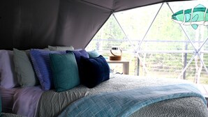 A bed with multiple pillows and a blanket, a small table with a basket, and a view of greenery through a window. - Unique Glamping Dome with Private Firepit Luxury Escape in Salt Lick, Kentucky (Salt Lick)