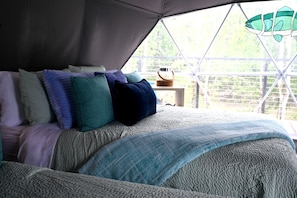Una cama con varias almohadas y una colcha, una mesita con una cesta, y vistas a la vegetación a través de una ventana.