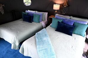 Habitación con dos camas, cada una con cubrecama blanco texturizado y varios cojines de distintos colores. Hay una mesita con una lámpara y adornos en la pared.