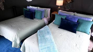 A room with two beds, each with a white textured bedspread and multiple pillows in various colors. There is a small table with a lamp and decorative items on the wall.
