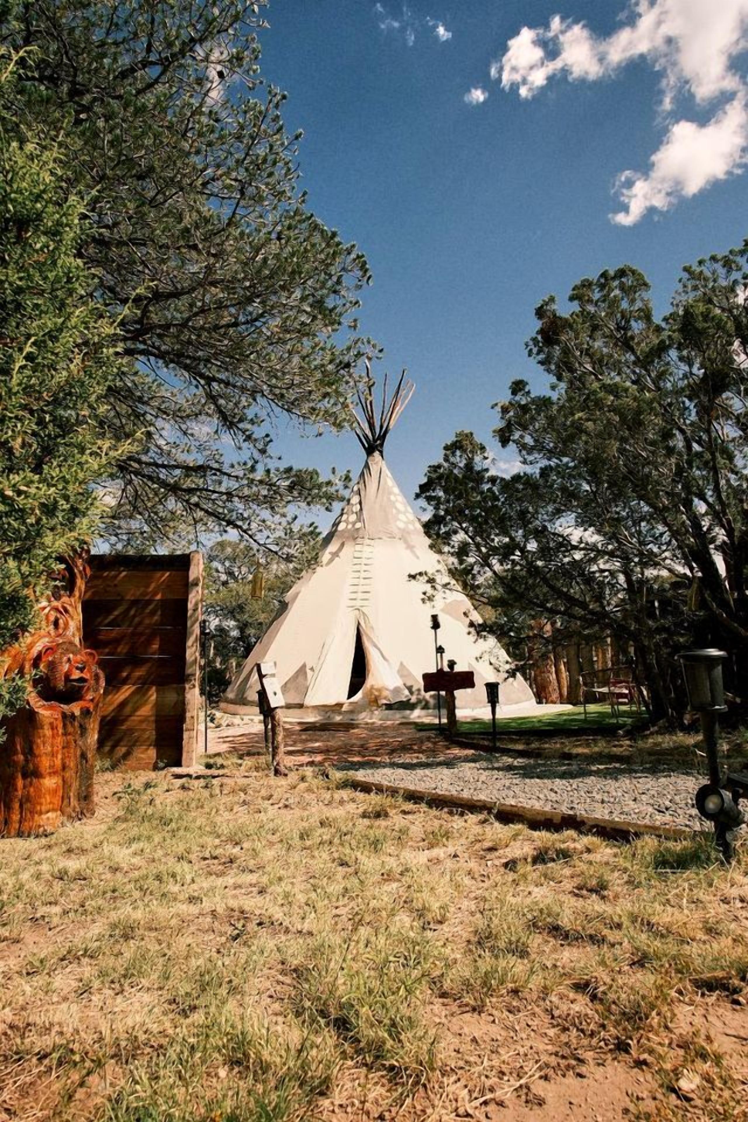 En traditionel tipi omgivet af træer, med et træhegn og en metal lygtepæl.