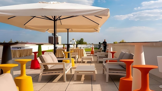 An outdoor seating area with white and orange furniture, a large umbrella, and a view of the sky. - Sports Academy Hotel (Balatonfured)