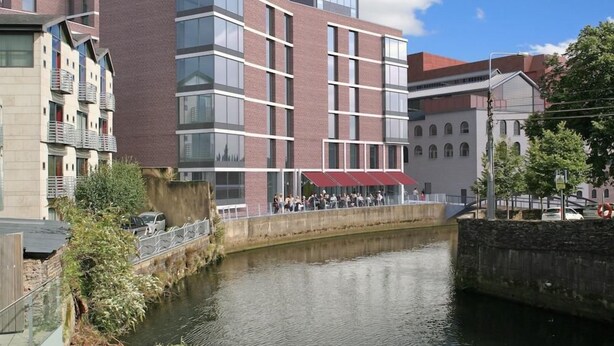 A modern building with a river in front, featuring a bridge and a red awning. - Yugo Lee Point (Cork)