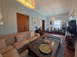 Ein Wohnzimmer mit einer Couch, einem Couchtisch und einem Fernseher. An den Wänden hängen gerahmte Bilder und es gibt eine Holztür.