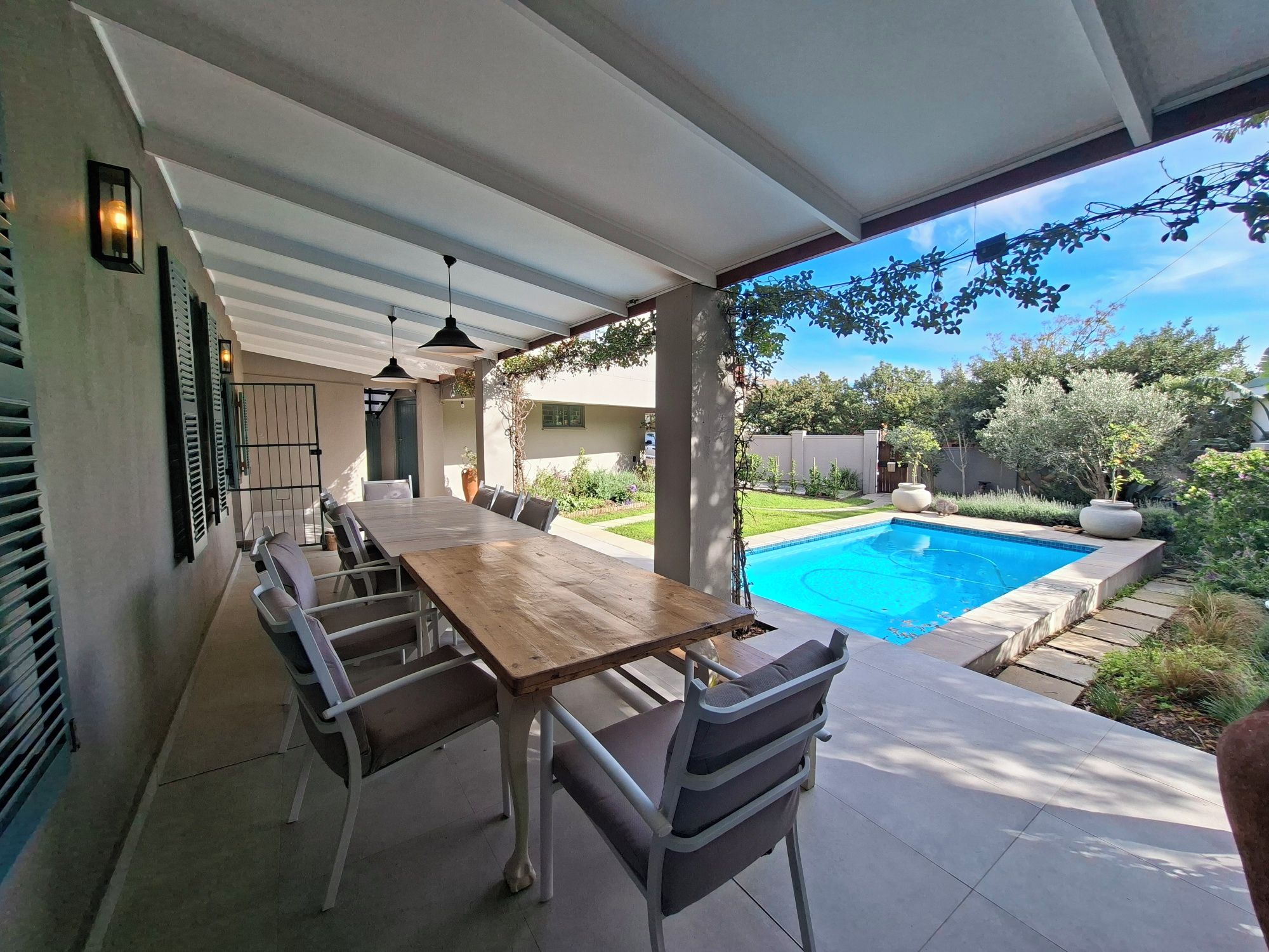 An outdoor patio with a wooden table and chairs, a swimming pool, and a covered area with hanging lights.