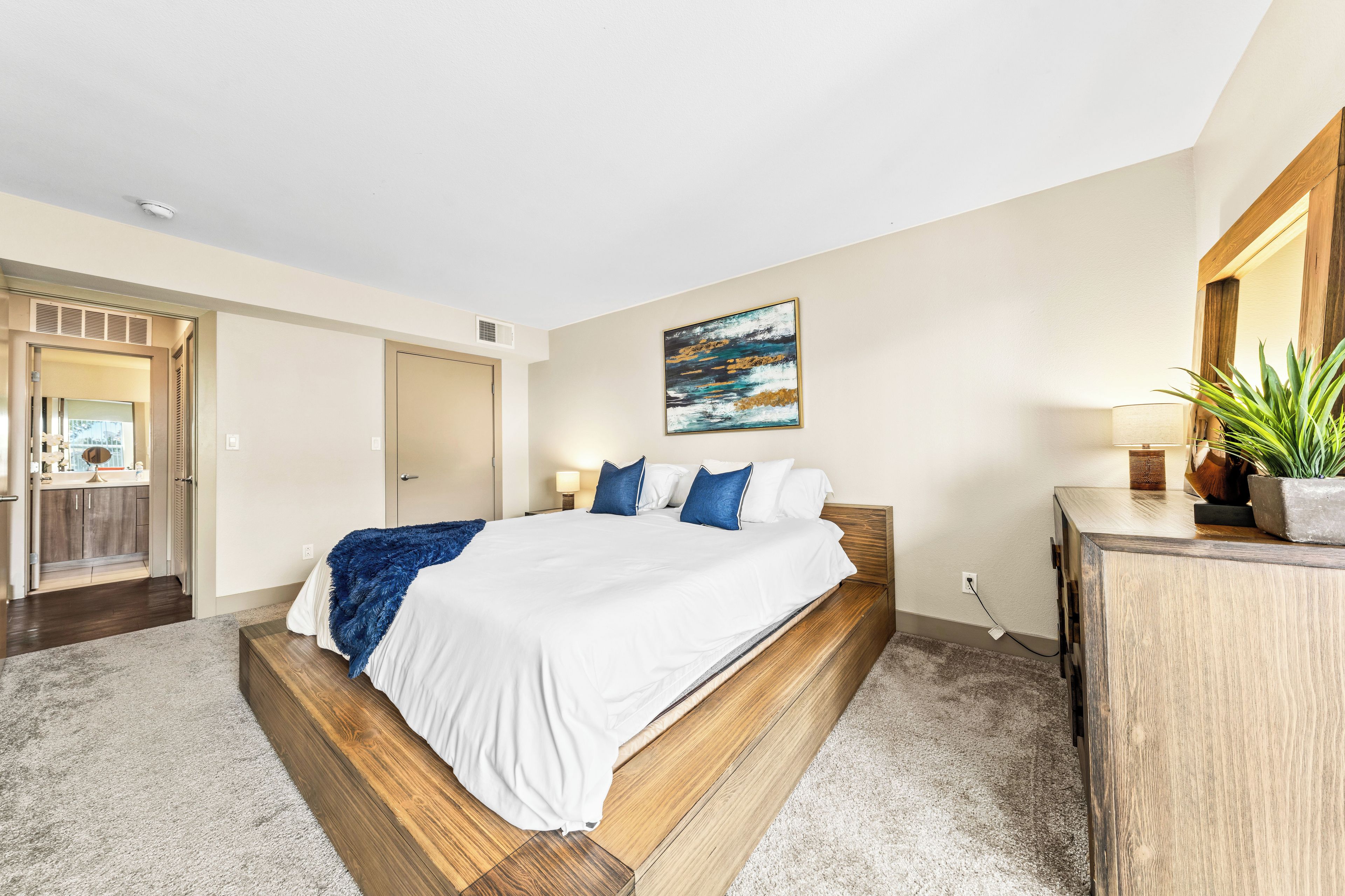 A hotel room with a large bed, a bedside table, a mirror, a painting on the wall, and a potted plant.